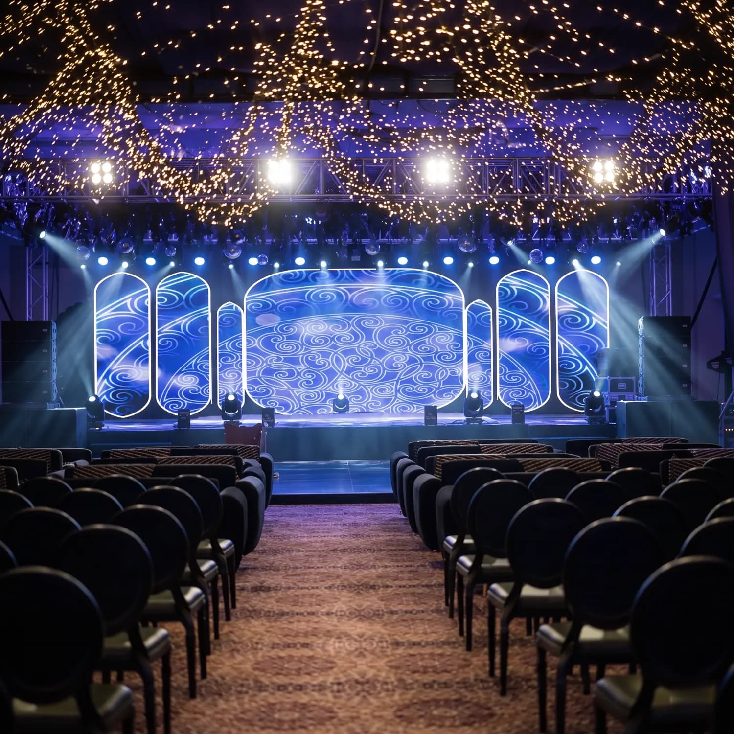 Stage with patterned LED screens, moving lights and a fairy-light ceiling above rows of seating